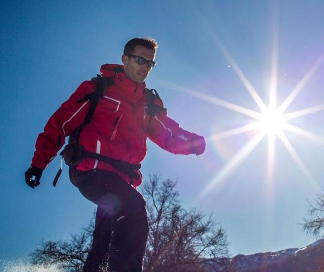 Homme en tenue de randonnée marchant sur la neige avec des raquettes, avec un soleil éclatant en arrière-plan.